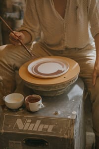 An artisan decorates a ceramic plate on a pottery wheel in an intimate workshop setting.